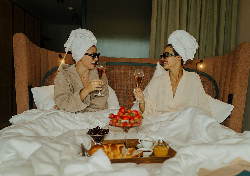 Zwei Frauen im Bademantel genießen ein Glas Sekt im Bett, mit einem Tablett voller Erdbeeren, Kirschen und Kuchen davor.