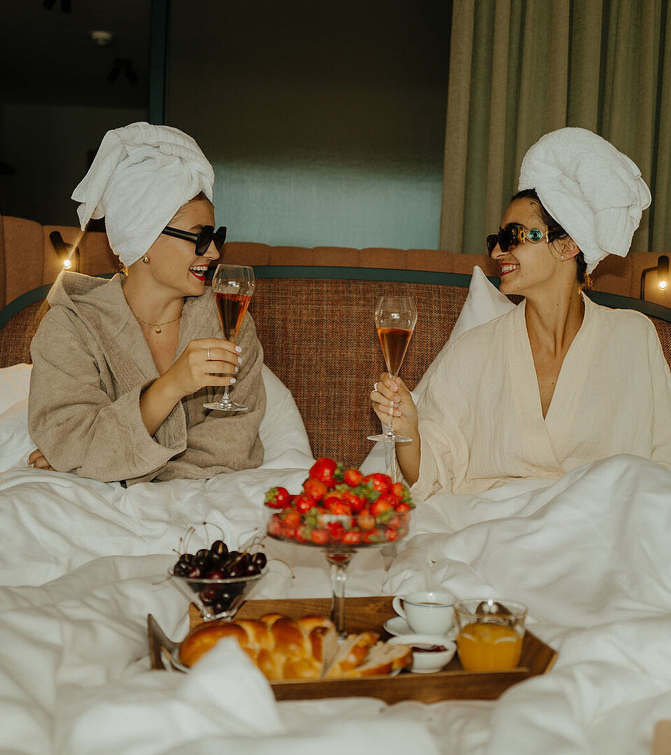 Zwei Frauen im Bademantel genießen ein Glas Sekt im Bett, mit einem Tablett voller Erdbeeren, Kirschen und Kuchen davor.