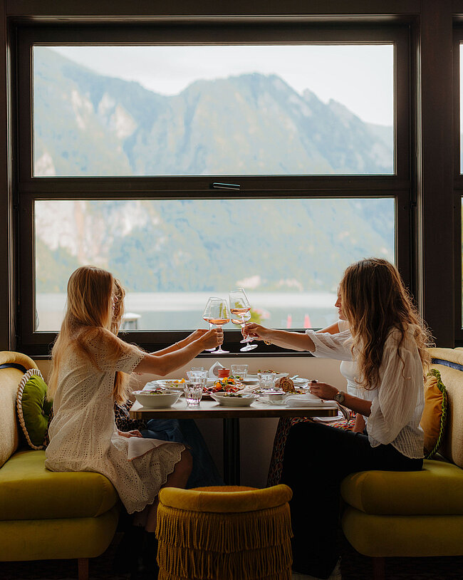 Vier Frauen genießen ein gemeinsames Abendessen, stoßen an und teilen sich Speisen, während im Hintergrund die Berge und der See sichtbar sind