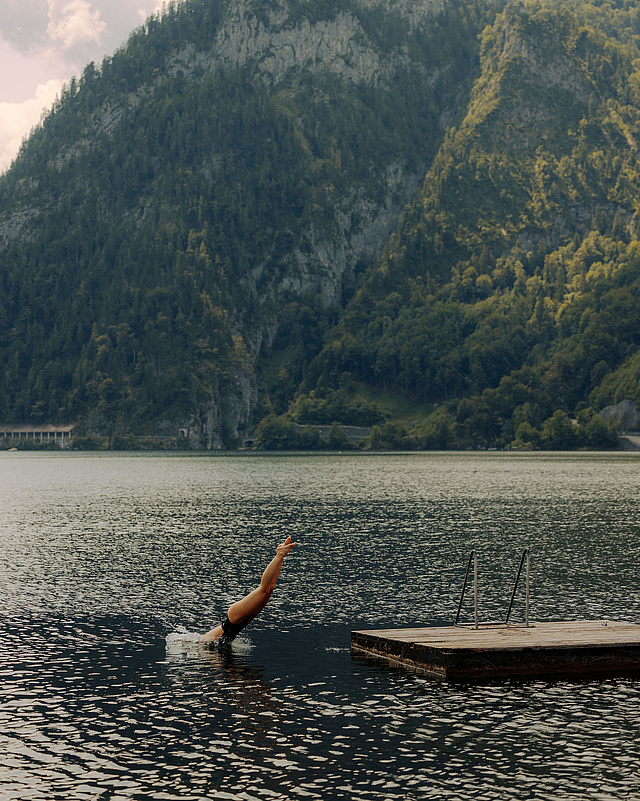 Person springt ins Wasser des Traunsees von einer Holzinsel aus, im Hintergrund Berge und ein klarer Sommerhimmel.