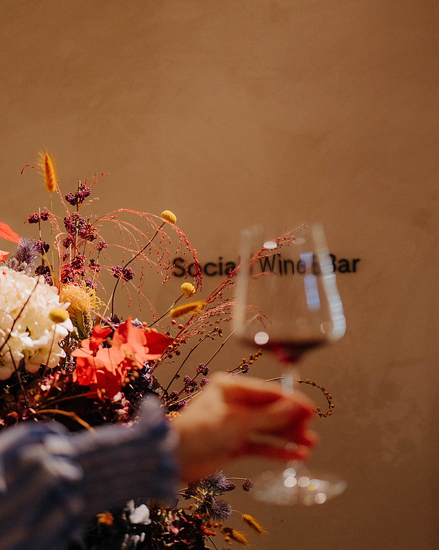 Verschwommene Hand mit Weinglas im Vordergrund, dahinter ein großes Blumenarrangement und der Schriftzug „Social Wine Bar“ an der Wand