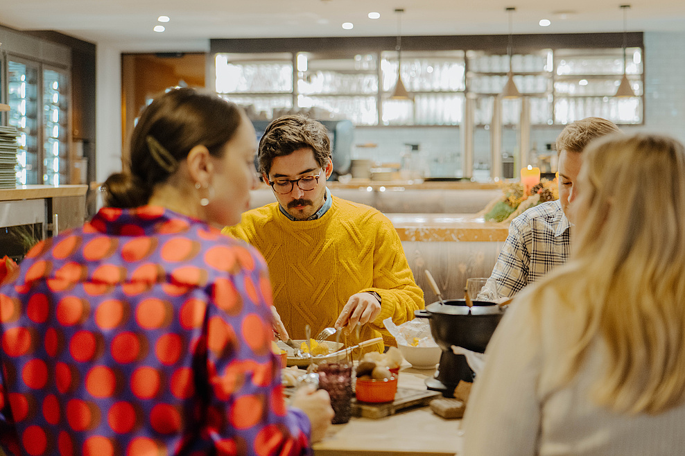 Vier Personen in bunter Kleidung genießen Fondue im Restaurant, im Hintergrund der Barbereich.