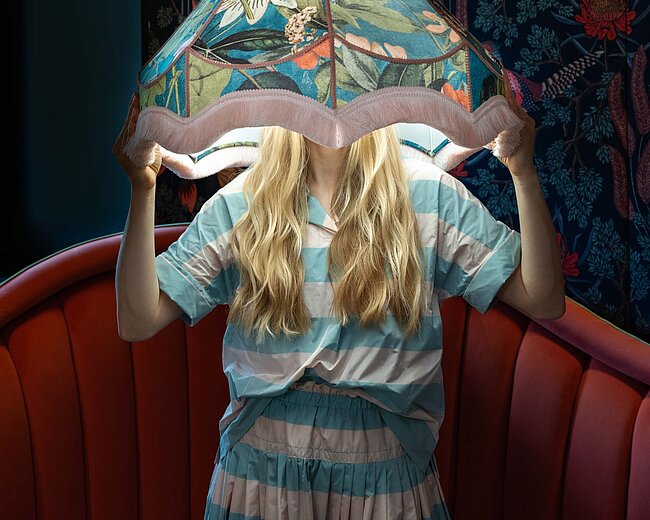 Model mit langen blonden Haaren, das einen bunten Lampenschirm trägt, vor einer roten Sitzecke und einer blumigen Tapete.